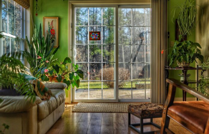 Sunlit living room with green walls, lush houseplants, and a large glass sliding door opening to a patio outside, with a small colorful stained-glass ornament on the door.