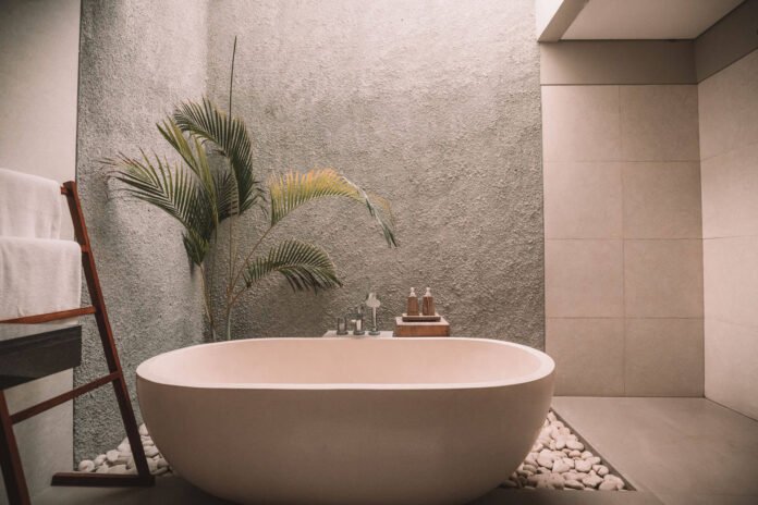 Freestanding oval bathtub in a modern spa-like bathroom with pebble floor and a palm plant nearby.