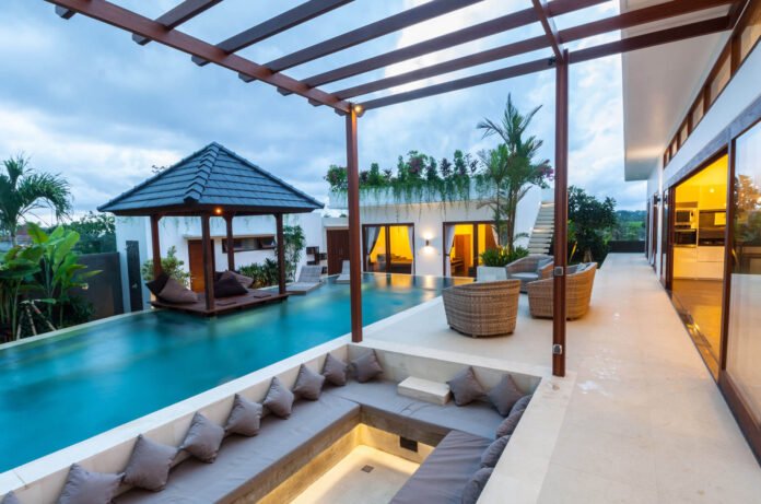 Luxurious outdoor pool area of a modern villa with a wooden pergola, lounge seating, and glass-walled interiors.