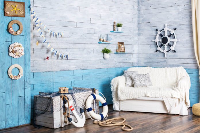 Cozy coastal room with blue weathered walls, a white sofa, striped cushions, and a white ship wheel on the wall.