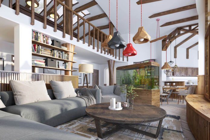 Spacious living area with a gray sectional sofa, round wooden coffee table, and a tall bookcase; exposed timber beams and colorful pendant lights hanging from the ceiling booting a modern rustic style.