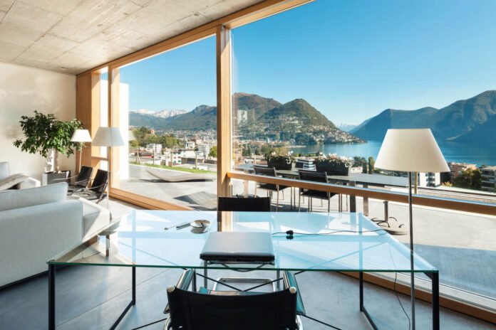Open-plan living area with floor-to-ceiling windows overlooking a lake and mountains, glass dining table and modern chairs.