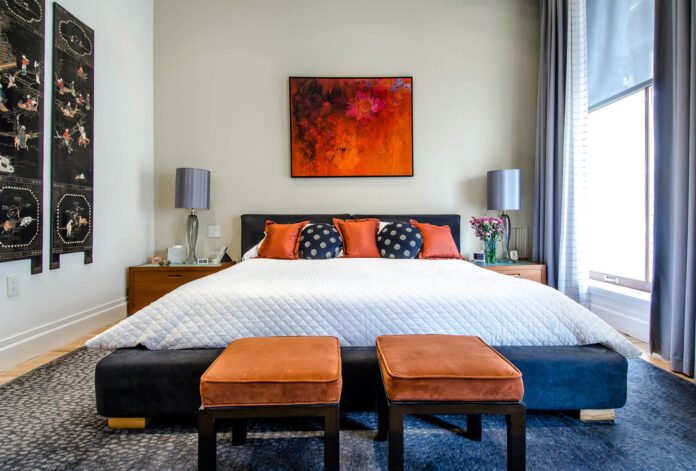Queen bed centered in a contemporary bedroom with orange-and-navy throw pillows, a white quilt, and three orange ottomans at the foot of the bed.