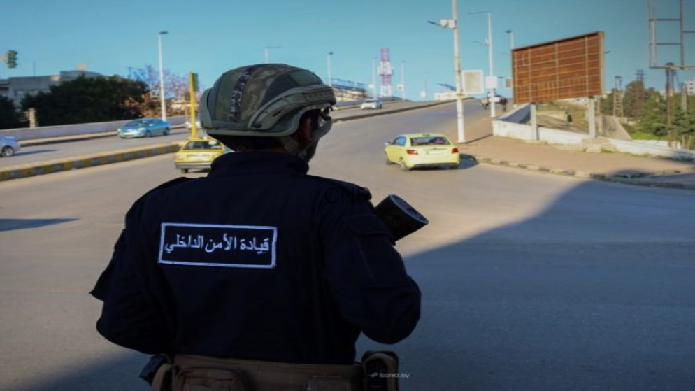 Security officer in helmet and dark uniform seen from behind, standing at a road junction with cars in the distance.
