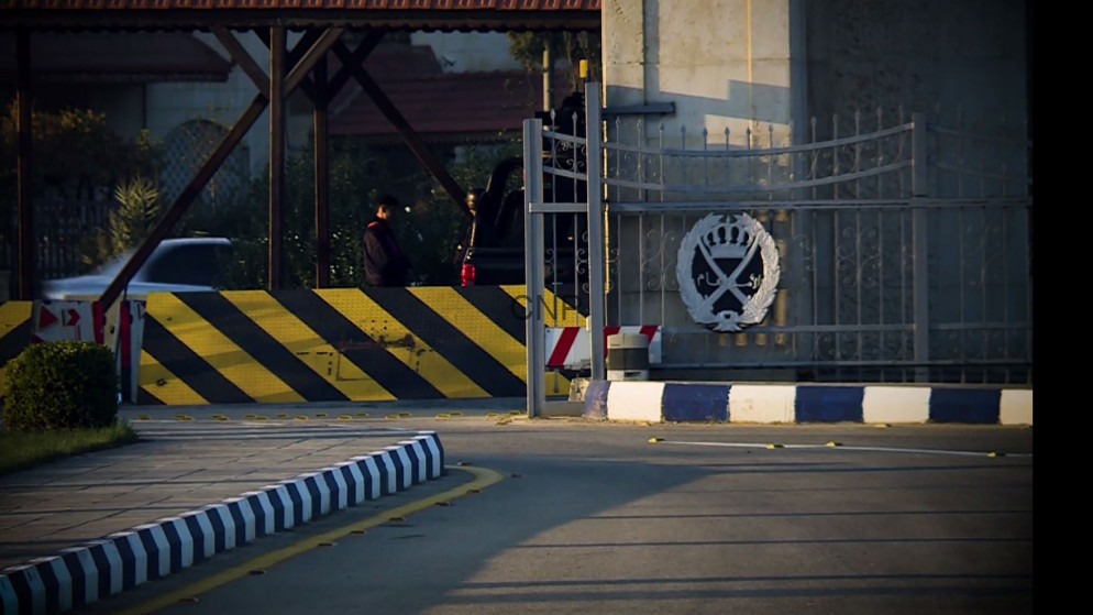 Gated security entrance with a decorative crest on the gate and a black-and-yellow striped barrier along a curved road, with a person in the background.