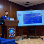 Male lecturer in a blue academic gown stands at a wooden podium with a university emblem, delivering a presentation as a large screen shows a blue slide.