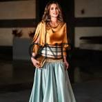image - Center News Network Woman in a Renaissance-style orange top and light blue skirt with a corset belt, standing in a dim room while holding a guitar.